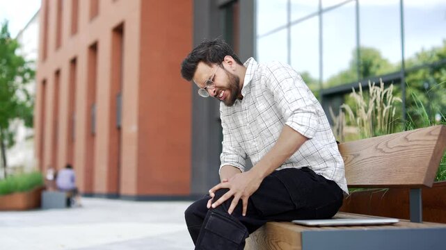 Young adult man suffers from painful severe knee joint pain sitting on a bench on street near a modern building. Upset bearded male massages leg muscles, could not walk, illness, cramps or rheumatism
