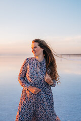 A young woman with long, windblown hair smiles peacefully by a calm lake during a golden sunset, radiating freedom, happiness, and natural beauty in a serene atmosphere.