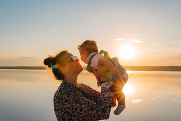 A loving mother lifting her happy baby during a warm sunset by a scenic lake, radiating joy and serenity.