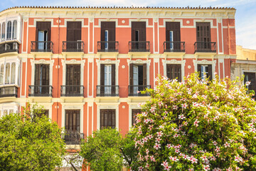 Fototapeta premium Malaga, seaside city in Andalusia, Spain, Europe. A peach-colored building with balconies and ornate details, partially obscured by lush green trees and flowering bushes.