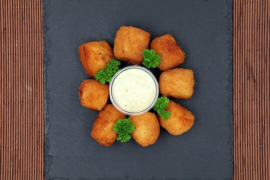 
Fried kibbeling with a jar of tartare sauce and parsley served on a slate. Fried pieces of cod fish in top view.
