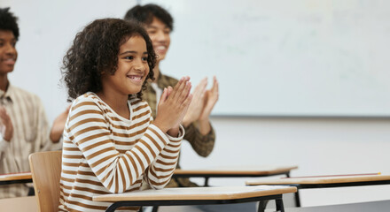 A girl in a striped shirt claps her hands while sitting at a desk in a classroom