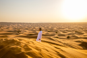 A man walks through the vast desert at golden hour, surrounded by the endless sand dunes of the Middle East. The warm sunlight highlights the peaceful, serene landscape.
