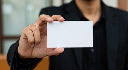 Fototapeta premium A close-up of a person holding a blank white business card at arm's length featuring a warm and inviting indoor setting the individual is wearing formal attire