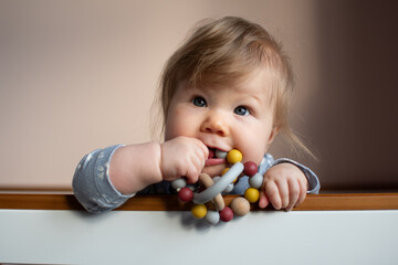 Cute Caucasian little baby girl standing in her crib and chewing on an infant toy, looking at the camera, with copy space. Funny baby