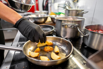 A chef in black gloves skillfully cooks diced potatoes in a stainless steel pan on a stovetop,...