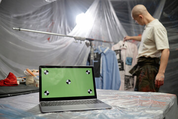 Open laptop displaying green mockup screen placed on working surface covered with masking film in fashion artists studio, copy space
