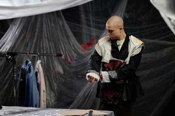 Medium full shot of adult male tailor with buzz cut trying on custom jacket creating eclectic patchwork design in workshop all covered with masking film, copy space