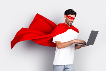 Cheerful man wearing a red superhero cape and mask using a laptop while standing against a white background © deagreez