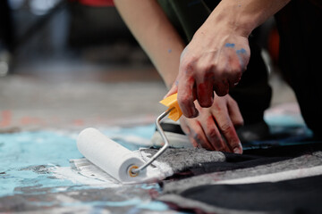 Close up on hands of male artist painting fabric with roller coloring in white custom made clothing item while working on contemporary fashion artwork on floor in studio, copy space