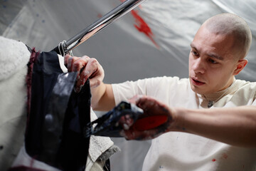 Dutch angle shot of concentrated male fashion artist fixating details on custom made clothes using...