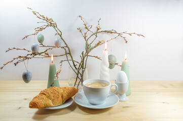 Coffee break with croissant and boiled egg, bouquet of spring branches with hanging blue gray Easter eggs and burning candles on a wooden table against a light wall, copy space