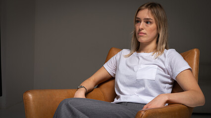 young, emotional, business girl sitting on a brown chair, wearing a white t-shirt, stress, worries, looking to the side. Modern minimalist portrait of a business or life theme, place for an inscriptio