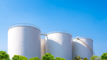 Storage fuel tanks behind row of green trees against blue sky background, Improving industrial landscape area with sustainable environmental concept, low angle view © Prapat