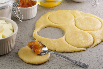 Preparation of a sweet pastry dough takes place in a cozy kitchen. Dough is rolled out and circular cutouts are combined with fruit filling and other baking ingredients.