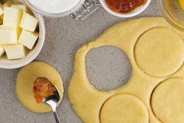 Preparation of a sweet pastry dough takes place in a cozy kitchen. Dough is rolled out and circular cutouts are combined with fruit filling and other baking ingredients.