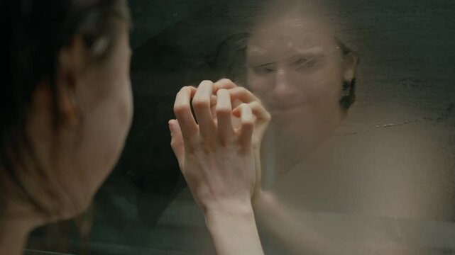 Over the shoulder view of emotionally unbalanced girl clearing foggy bathroom mirror with palm and hitting it with fist while looking at her reflection