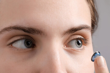 Woman putting in blue color contact lens on grey background, closeup
