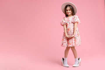 Stylish little girl wearing oversized high heeled shoes on pink background, space for text