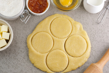 Freshly rolled dough is placed on a gray countertop, featuring circular cutouts ready for baking. Nearby, bowls of flour, butter, jam, and an egg are visible, setting the stage for pastry making.