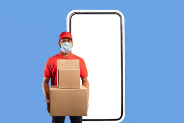 Delivery Application. African American Courier Standing Near Big Phone Screen Holding Cardboard Boxes Looking At Camera Over Yellow Studio Background, Wearing Face Mask. Square
