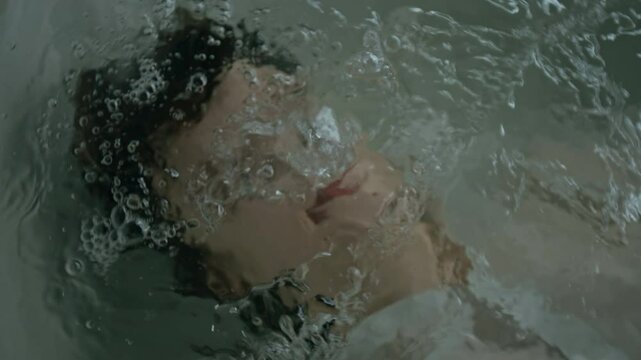 Close-up view of stressed young woman going under water in bathtub with closed eyes, then sweeping hands across her face