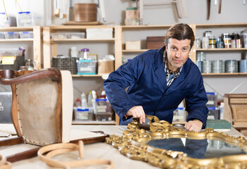 Skilled middle-aged male furniture repair specialist in uniform restoring Victorian mirror frame during workday in workshop