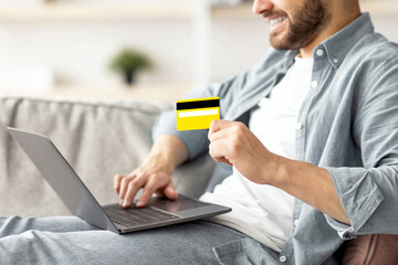 Online shopping. Cropped view of young guy with laptop and credit card sitting on couch at home, purchasing goods in web store. Unrecognizable man buying things on internet, using contactless payment