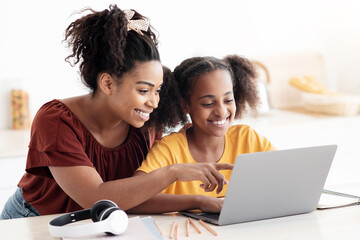 Creative black mother and daughter working on school project, using modern laptop, taking notes, cheerful afro-american woman helping her teen kid with homework, kitchen table, copy space