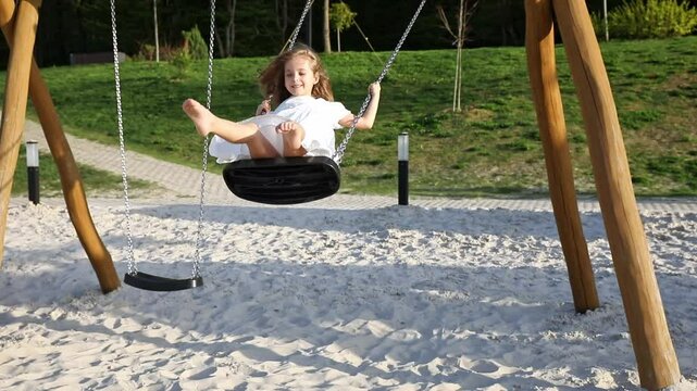 Happy joyful little girl in white summer dress rides on a swing on a sandy beach with green lawn background or on playground in children park, on a sunny day. Vacation concept. Children's Day footage