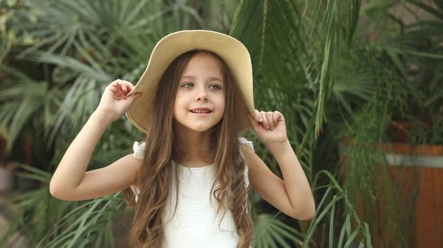 Cute happy little girl in a white summer dress and straw hat near palm leaves. Child in the botanical garden or greenhouse. Tropical vacation concept. Footage