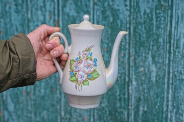hand holding one white ceramic teapot with colorful pattern on green wall background