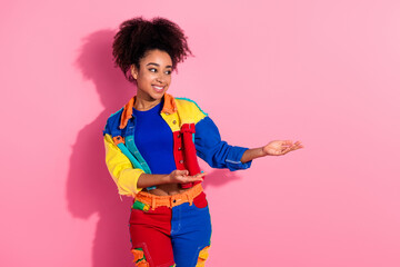 Stylish cheerful woman standing against pink background with vibrant multicolored outfit and joyful gesture of presenting or showing.