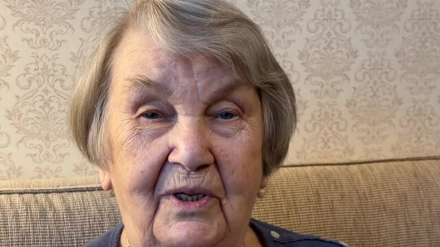 The face and eyes of an elderly woman of the 80s with gray hair, smiling happily, with wrinkled skin, who is sitting on the couch, looking at the camera and talking about something. Close-up.