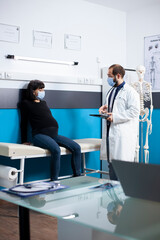 Young doctor in medical office consults pregnant patient about her health, using tablet to discuss upcoming maternity plan. Male gynecologist provides expectant woman with pregnancy treatment options.