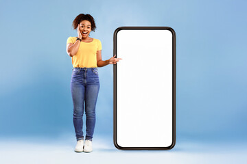 Emotional African American woman cannot believe great offer, pointing at big smartphone with space for new mobile app on screen, blue studio background. Mockup for website. Cellphone display template