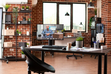 Vacant brick wall workplace setup with computer screen showing videocall of businessman in wheelchair. Desktop pc displaying ongoing virtual meeting with caucasian man in empty startup office.