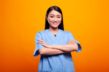 Cheerful young female doctor wearing blue scrubs is confidently posing with her arms positioned...