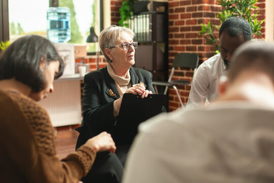Caucasian retired female psychiatrist holding clipboard and giving advice to multiethnic clients during therapy session. Elderly woman psychologist providing guidance to diverse group at aa meeting.