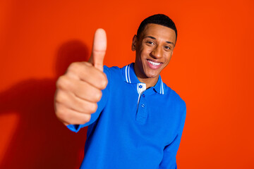 Smiling young man in blue polo giving thumbs up on orange background