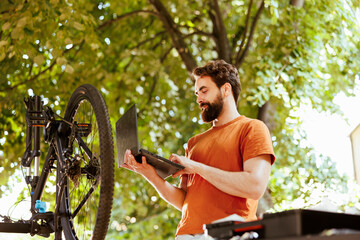 Athletic caucasian male cyclist utilizing minicomputer for bike maintanence research. Healthy dedicated young man engrossed in inspecting and servicing modern bicycle with laptop.