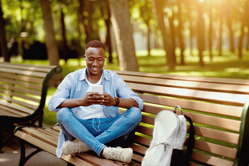 Obraz premium Funky black guy in earphones listening to his favorite music online from cellphone on bench at park, free space