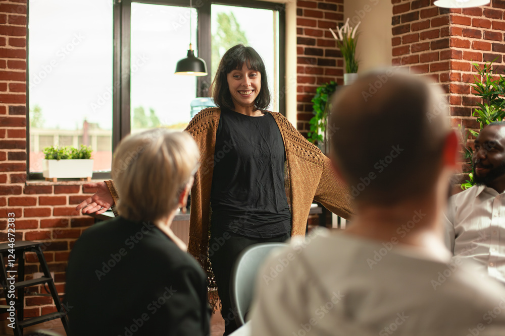 Wall mural Smiling white woman standing in circle of people, discussing her progress at therapy session in supportive space. Young female patient excitedly shares her recovery achievement at aa group meeting.