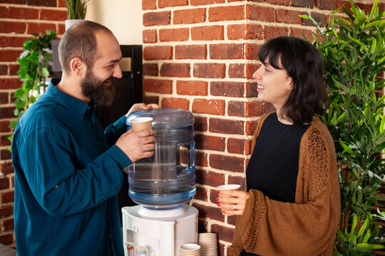 Smiling caucasian employees hold coffee cups, engaging in casual yet productive conversation in brick wall office. Businessman and woman having relaxed interaction with refreshment near water cooler. - Powered by Adobe