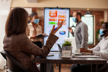 Multiethnic team wearing face masks for safety in office and discussing strategy ideas with information seen on monitor. Closeup on woman with raised hand, holding a pen and speaking to her coworkers.