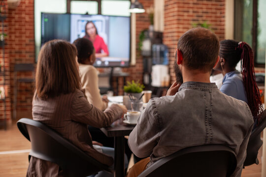 Bearded man and coworkers discuss marketing plan, watching digital screen displaying female client on video call. Diverse team conduct business meeting with remote businesswoman visible on monitor.