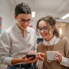 two female colleagues on the coffee break use mobile phone at office