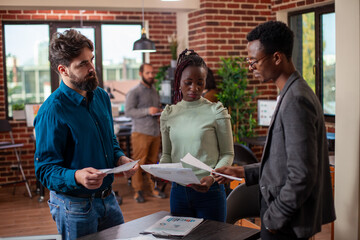 Startup employees stand around a table, reviewing company growth ideas and analyzing data in collaborative setting. Male and female colleagues holding documents and brainstorming strategies in office.