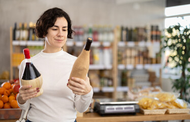 Woman choosing compare varieties red wine, doubt and think before buying. Client choose and decide purchase bottle of red wine, inspect good. Shop visitor pick up food in minimarket