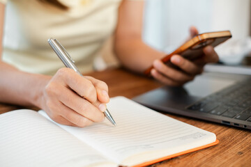 Close-up of a person writing in a notebook while holding a smartphone, with a laptop nearby on a wooden desk.